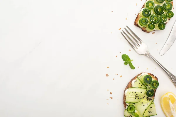 Vista superior de tostadas frescas de pepino con semillas, hojas de albahaca y cubiertos sobre fondo blanco - foto de stock