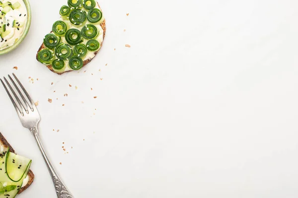 Вид зверху на свіжі тости огірків з насінням біля йогурту в мисці і виделкою на білому тлі — стокове фото