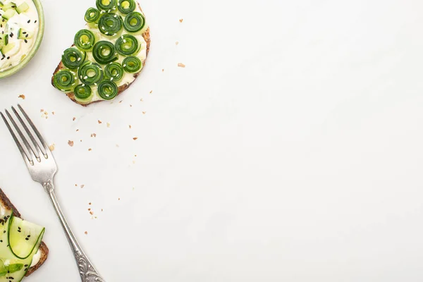 Top view of fresh cucumber toasts with seeds near yogurt in bowl and fork on white background — Stock Photo