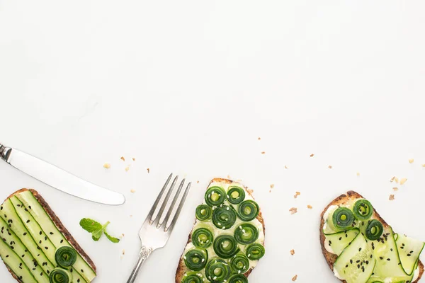 Вид на свежие огуречные тосты с семенами, мятными листьями и столовыми приборами на белом фоне — стоковое фото