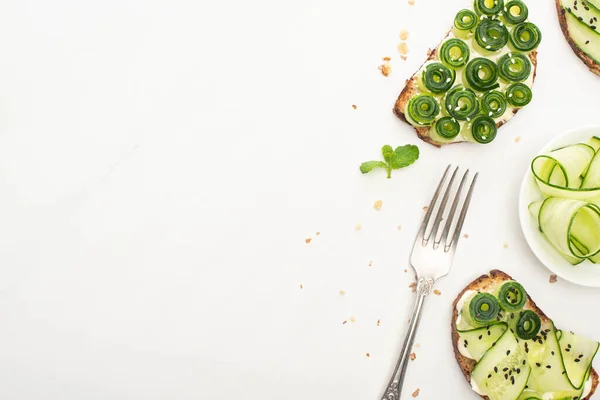 Vista superior de tostadas frescas de pepino con semillas, hojas de menta y tenedor sobre fondo blanco - foto de stock