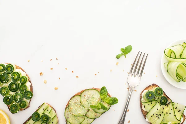 Vista superior de tostadas frescas de pepino con semillas, hojas de menta y albahaca y tenedor sobre fondo blanco - foto de stock