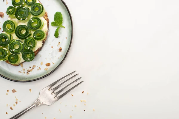 Vista superior de la tostada de pepino fresco con hojas de sésamo y menta en el plato cerca de tenedor sobre fondo blanco - foto de stock