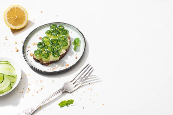 Pane tostato di cetriolo fresco con foglie di sesamo e menta sul piatto vicino a forchetta e limone su sfondo bianco — Foto stock