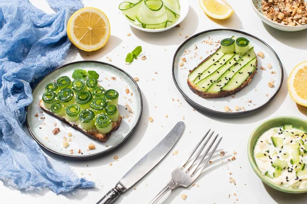 Fresh cucumber toasts on plates near cutlery, yogurt and lemon on white background — Stock Photo