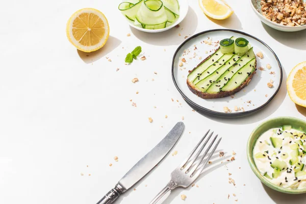 Tostadas de pepino fresco con semillas cerca de hojas de menta, cubiertos, yogur y limón sobre fondo blanco - foto de stock