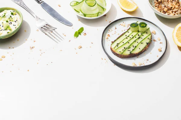 Tostadas de pepino fresco con semillas cerca de hojas de menta, cubiertos, yogur y limón sobre fondo blanco - foto de stock