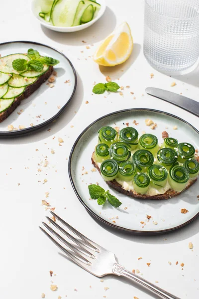 Fresh cucumber toasts with seeds, mint leaves and lemon near glass of water and cutlery on white background — Stock Photo