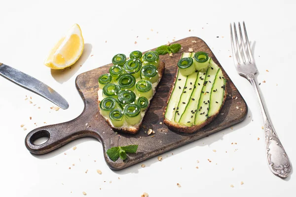 Torradas de pepino fresco em tábua de corte de madeira perto de talheres e limão no fundo branco — Fotografia de Stock