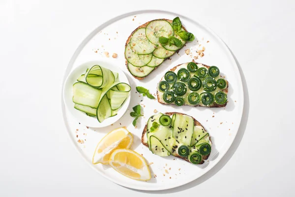 Top view of fresh cucumber toasts with seeds, mint and basil leaves on plate with lemon on white background — Stock Photo