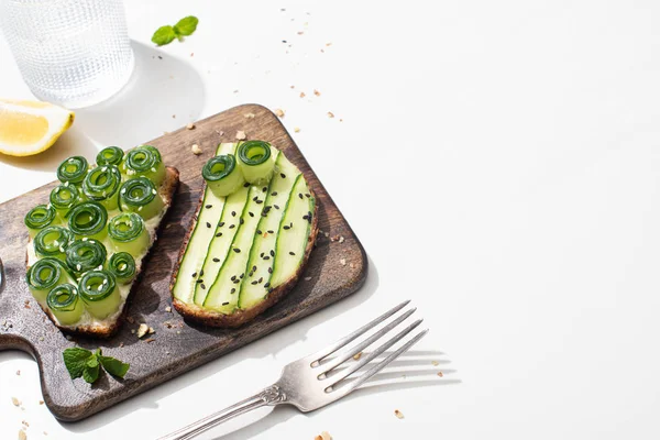 Pain grillé au concombre frais sur planche à découper en bois près de l'eau, fourchette et citron sur fond blanc — Photo de stock