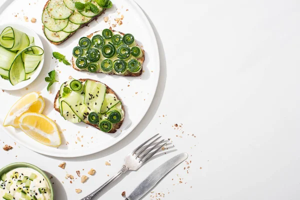 Top view of fresh cucumber toasts with seeds, mint and basil leaves on plate with lemon near cutlery and yogurt on white background — Stock Photo