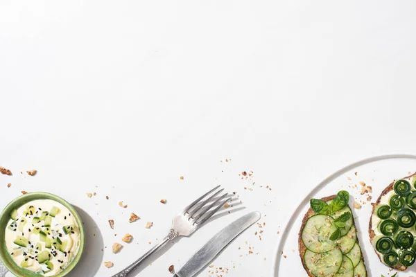 Vue de dessus des toasts au concombre frais avec des graines, de la menthe et des feuilles de basilic dans une assiette près des couverts et du yaourt sur fond blanc — Photo de stock