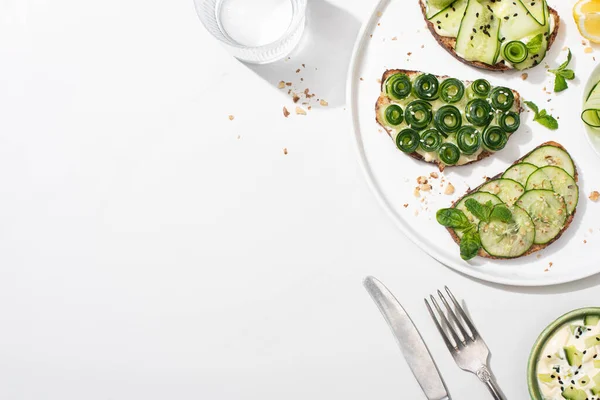 Top view of fresh cucumber toasts with seeds, mint and basil leaves on plate near cutlery, water and yogurt on white background — Stock Photo