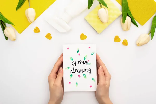 Vista cortada da mulher segurando primavera cartão de limpeza perto de tulipas primavera, suprimentos de limpeza amarelos no fundo branco — Fotografia de Stock