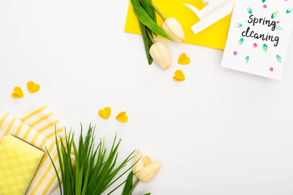 Vue de dessus des tulipes de printemps et de l'herbe verte près des fournitures de nettoyage jaunes et de la carte de nettoyage de printemps sur fond blanc — Photo de stock