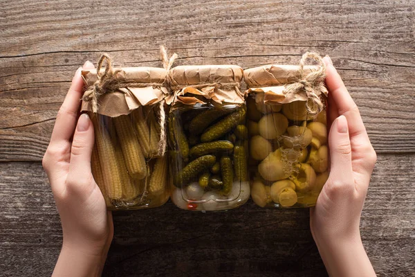 Cropped view of woman holding jars with delicious homemade tasty pickled cucumbers, corn and mushrooms on wooden rustic table — Stock Photo