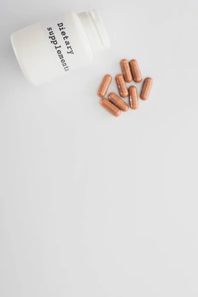 Top view of container with dietary supplements lettering and brown capsules on white background — Stock Photo