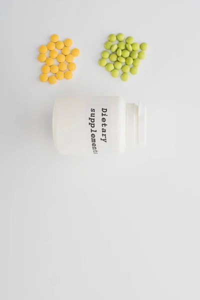 Top view of container with dietary supplements lettering and colorful pills on white background — Stock Photo