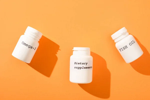 Top view of containers with omega-3, fish oil and dietary supplements lettering on orange background — Stock Photo