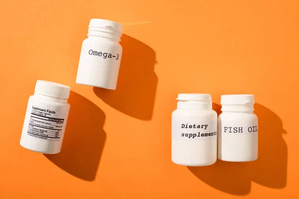 Top view of containers with omega-3, fish oil and dietary supplements lettering on orange background — Stock Photo