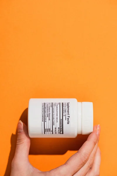 Partial view of woman holding white container with dietary supplements on orange background — Stock Photo