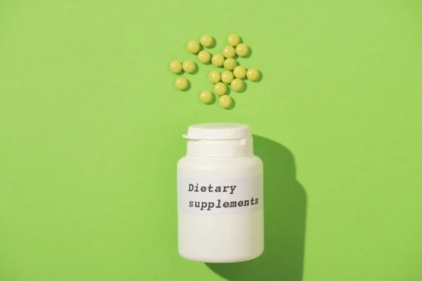 Top view of container with dietary supplements lettering and pills on green background — Stock Photo