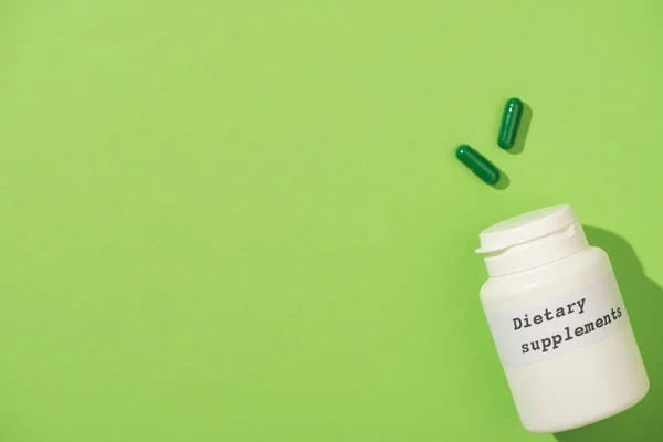 Top view of container with dietary supplements lettering and capsules on green — Stock Photo