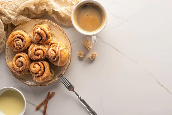 Vista dall'alto di rotoli di cannella freschi fatti in casa sulla superficie di marmo con tazza di caffè, latte condensato, forchetta e stoffa — Foto stock