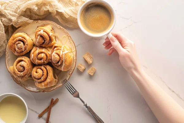 Cropped view of woman holding cup of coffee near homemade cinnamon rolls on marble surface with fork, condensed milk and cloth — Stock Photo