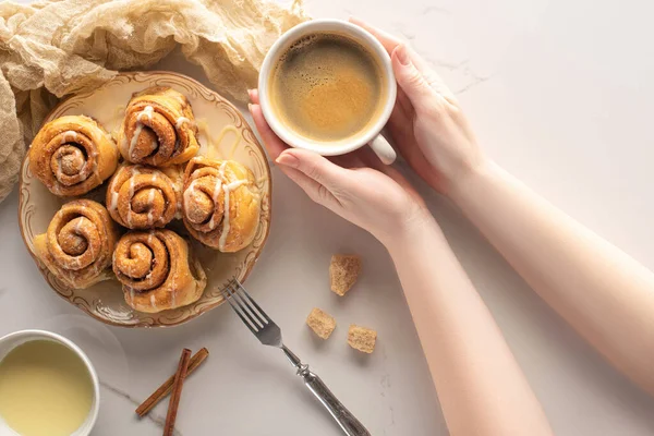 Cropped view of woman holding cup of coffee near homemade cinnamon rolls on marble surface with fork, condensed milk and cloth — Stock Photo