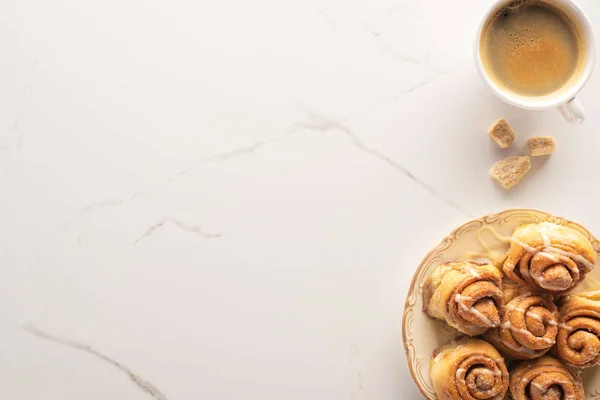 Vista superior de rollos de canela caseros frescos en la superficie de mármol con taza de café, azúcar morena - foto de stock