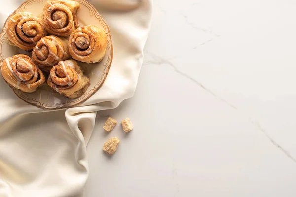 Vue de dessus des rouleaux de cannelle maison fraîche sur tissu de soie sur la surface de marbre avec sucre brun — Photo de stock