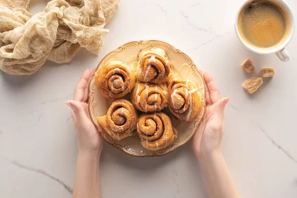 Cropped view of woman holding plate with fresh homemade cinnamon rolls on marble surface with cup of coffee and cloth — Stock Photo