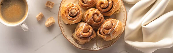 Top view of fresh homemade cinnamon rolls on marble surface with cup of coffee, condensed milk and silk cloth, panoramic shot — Stock Photo