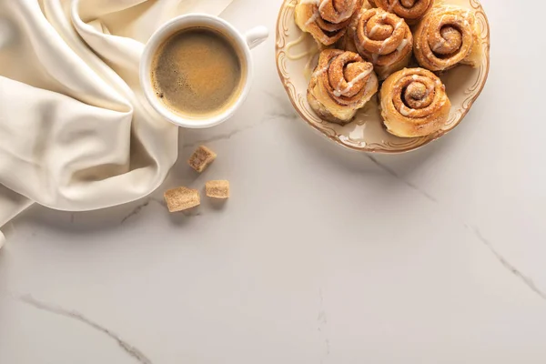 Vista superior de rolos de canela caseiros frescos na superfície de mármore com xícara de café, açúcar mascavo e pano de seda — Fotografia de Stock