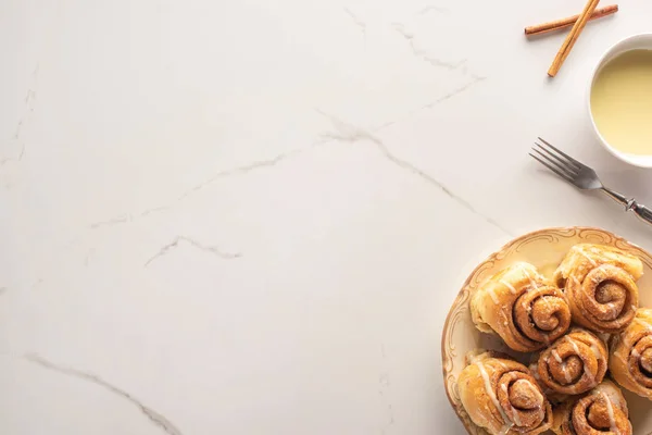 Vue du dessus des rouleaux de cannelle frais faits maison sur la surface du marbre avec du lait condensé, fourchette — Photo de stock