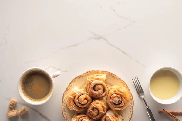 Vista superior de rolos de canela caseiros frescos na superfície de mármore com xícara de café, leite condensado, garfo — Fotografia de Stock