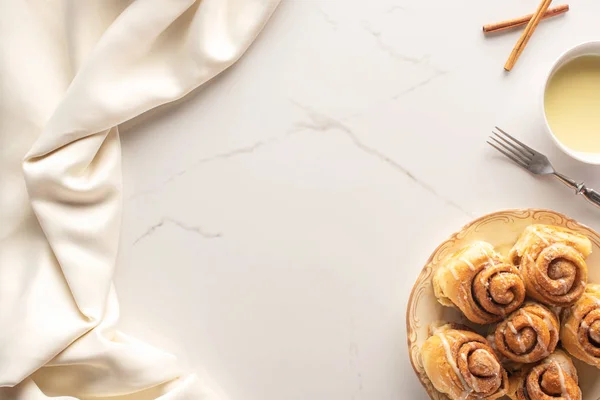 Vista superior de rolos de canela caseiros frescos na superfície de mármore com leite condensado, garfo e pano de cetim — Fotografia de Stock