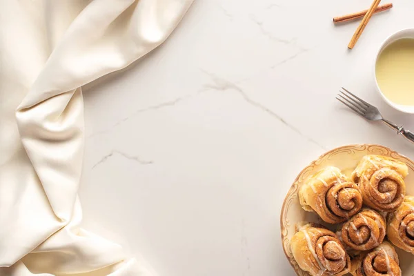 Vista dall'alto di rotoli di cannella freschi fatti in casa su superficie di marmo con latte condensato, forchetta e panno di raso — Foto stock