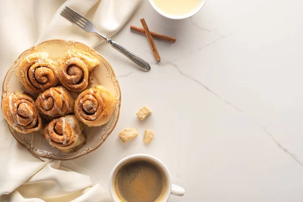 Draufsicht auf frische hausgemachte Zimtrollen auf Marmoroberfläche mit Kaffee, Kondensmilch, Gabel und Satintuch — Stockfoto