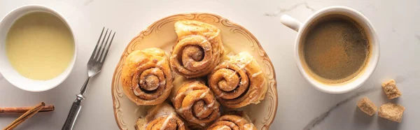 Vue de dessus des rouleaux de cannelle maison fraîche sur la surface du marbre avec tasse de café, fourchette et lait condensé, vue panoramique — Photo de stock