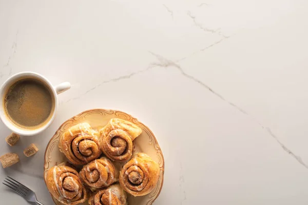 Vista dall'alto di rotoli di cannella freschi fatti in casa sulla superficie di marmo con tazza di caffè, forchetta e zucchero di canna — Foto stock