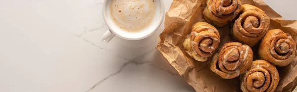 Top view of fresh homemade cinnamon rolls on parchment paper on marble surface with cup of coffee, panoramic shot — Stock Photo