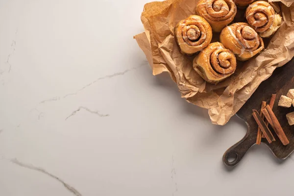 Vista dall'alto di rotoli di cannella freschi fatti in casa su carta pergamena su superficie di marmo con tagliere con zucchero di canna e bastoncini di cannella — Foto stock