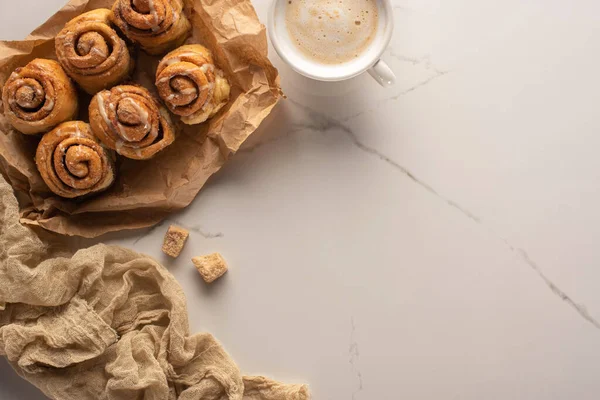 Vista superior de rollos de canela caseros frescos en papel pergamino sobre superficie de mármol con taza de café, azúcar morena y tela - foto de stock