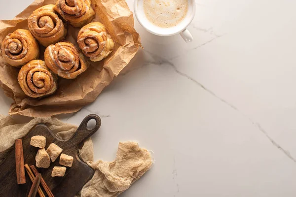Top view of fresh homemade cinnamon rolls on parchment paper on marble surface with cup of coffee and cutting board with brown sugar and cinnamon sticks on cloth — Stock Photo