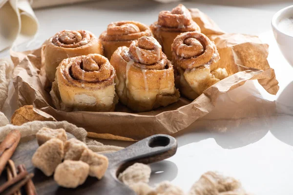 Enfoque selectivo de rollos de canela caseros frescos cerca de la tabla de cortar con azúcar morena y palitos de canela — Stock Photo