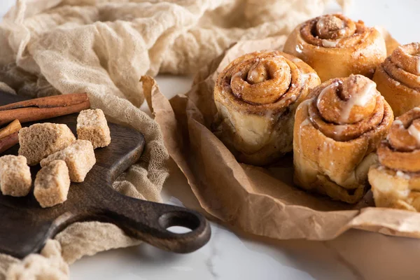 Selective focus of fresh homemade cinnamon rolls near cutting board with brown sugar and cinnamon sticks — Stock Photo