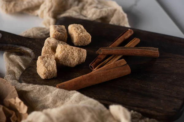 Bâtonnets de cannelle frais et sucre brun sur planche à découper en bois près du tissu — Photo de stock